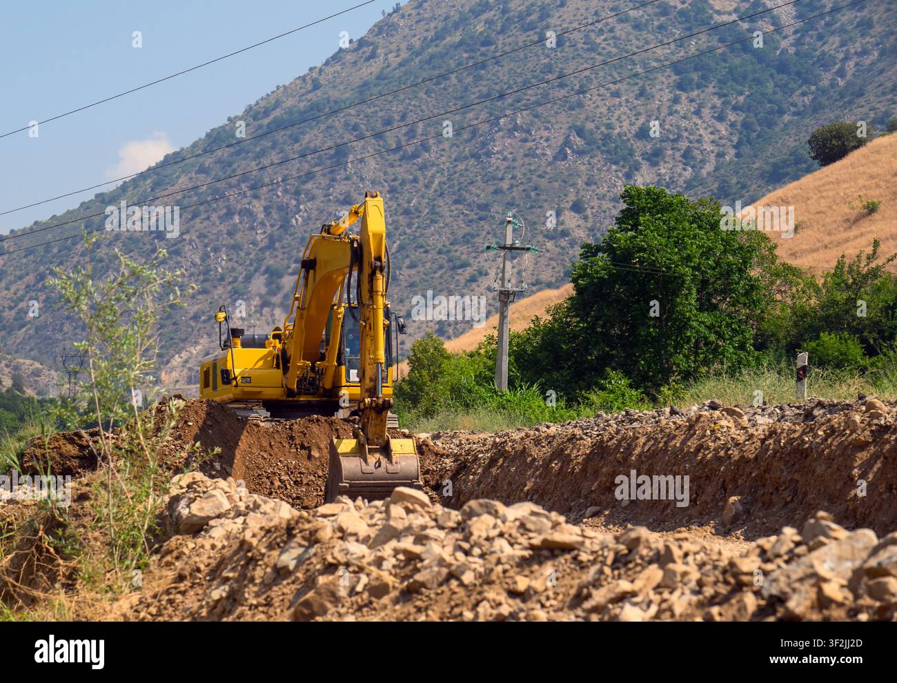 Excavateur jaune creuse un fossé dans un terrain rocheux. Fond montagneux avec des arbres verts Banque D'Images
