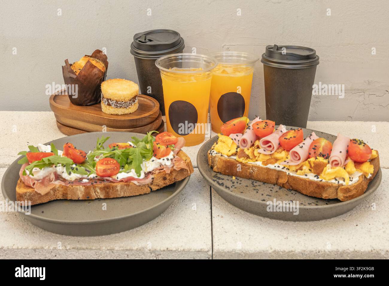 Deux toasts savoureux garnis d'œufs, de jambon, de tomates et d'arugula servis avec du jus d'orange, du café et des pâtisseries sur une surface en béton gris. Banque D'Images