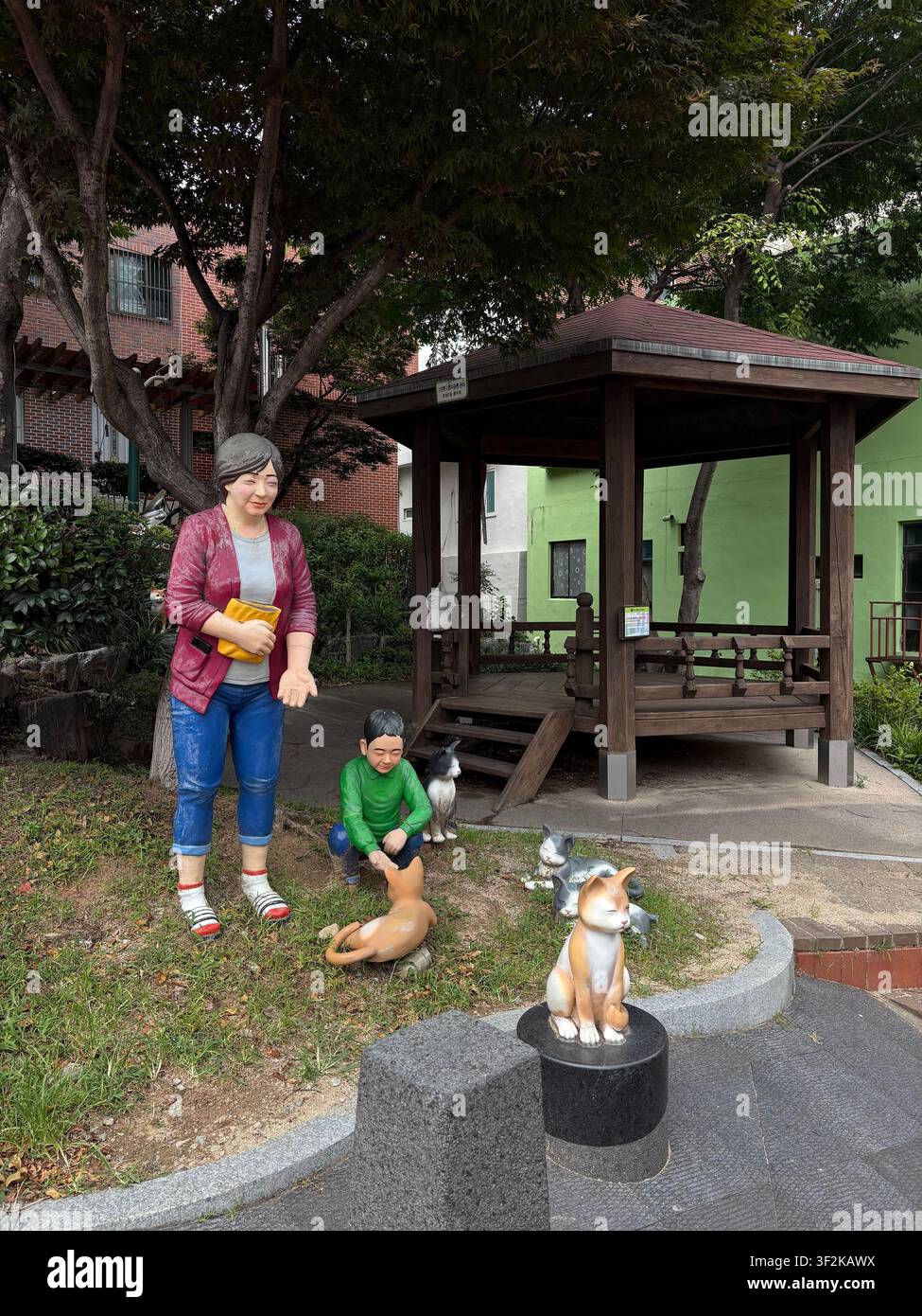 15 juillet 2025, Busan, Corée du Sud – Figurines peintes grandeur nature de personnes et de chiens exposées dans une rue résidentielle calme, avec une structure de porte en bois traditionnelle. - Image de stock capturée avec un smartphone