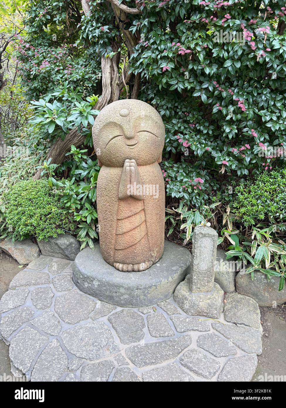 3 octobre 2025, Kamakura, Japon – Vue aérienne d’une statue de Jizo Bosatsu en pierre érodée reposant parmi des plantes de jardin vertes luxuriantes et un chemin de pierre. - Image de stock capturée avec un smartphone