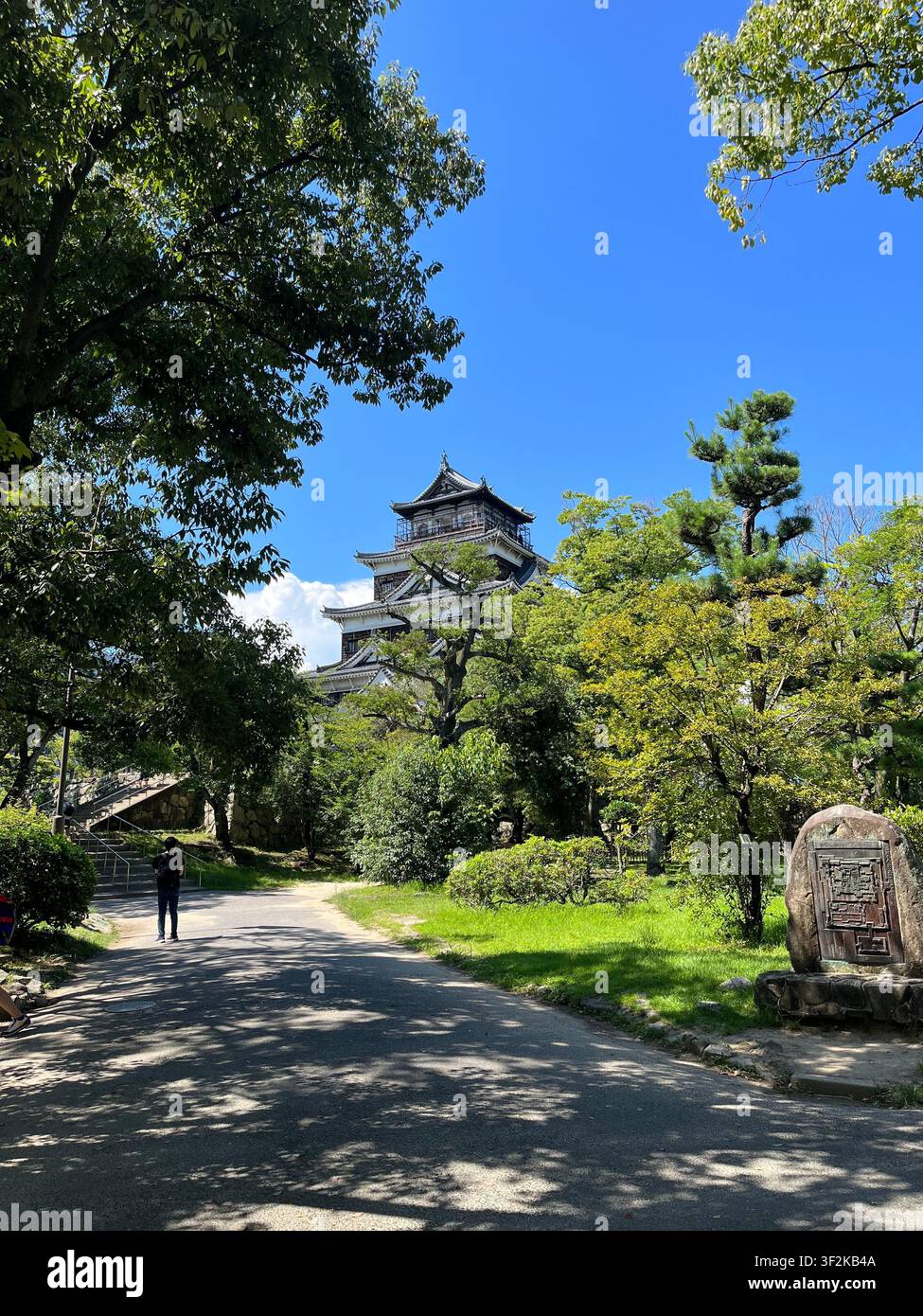25 juillet 2023, Hiroshima, Japon – La tour historique du château d’Hiroshima s’élève au-dessus des arbres verdoyants de l’été sous un ciel bleu, vue depuis un sentier des terrains du château. Banque D'Images
