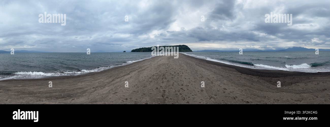 Vue panoramique dramatique d'une langue de sable noir volcanique s'étendant vers une île verte dans la baie de Kagoshima, Ibusuki, Kyushu, Japon, sous des nuages orageux. - Image de stock capturée avec un smartphone