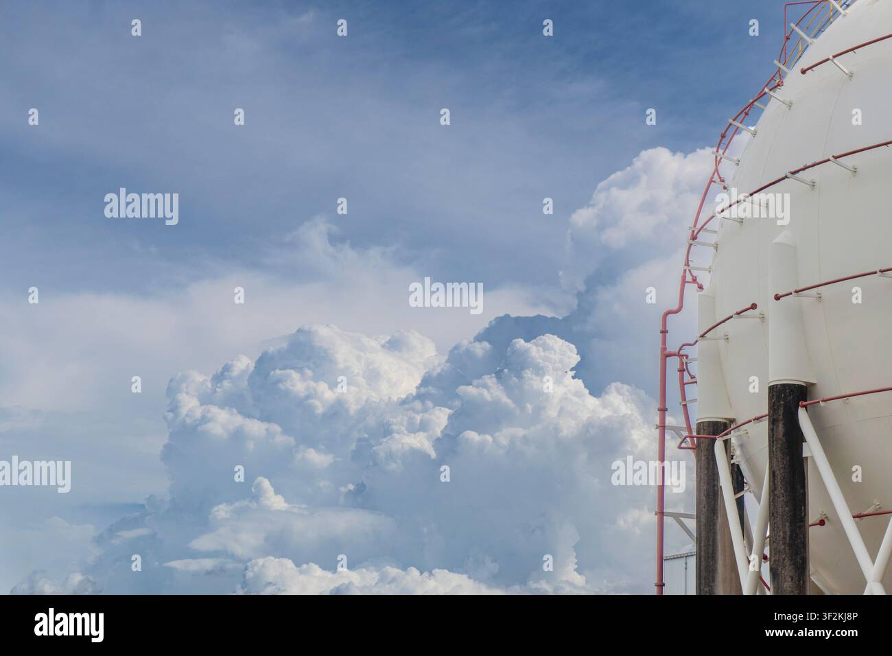Vue latérale d'un grand réservoir sphérique de gaz LPG avec des pipelines rouges et un escalier contre un ciel bleu dramatique avec des nuages cumulus imposants, capturant Banque D'Images