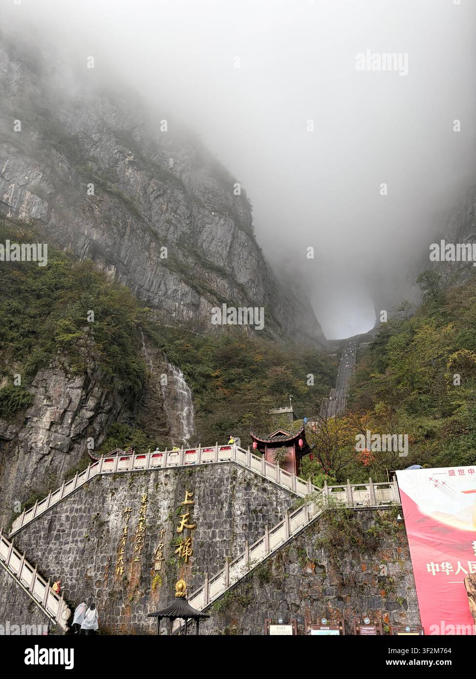 3 novembre 2025, Zhangjiajie, Chine – Un brouillard dramatique enveloppe des falaises de grès abruptes avec un escalier en pierre traditionnel, des inscriptions chinoises dorées et un pavillon dans la zone pittoresque de la montagne Tianmen. - Image de stock capturée avec un smartphone