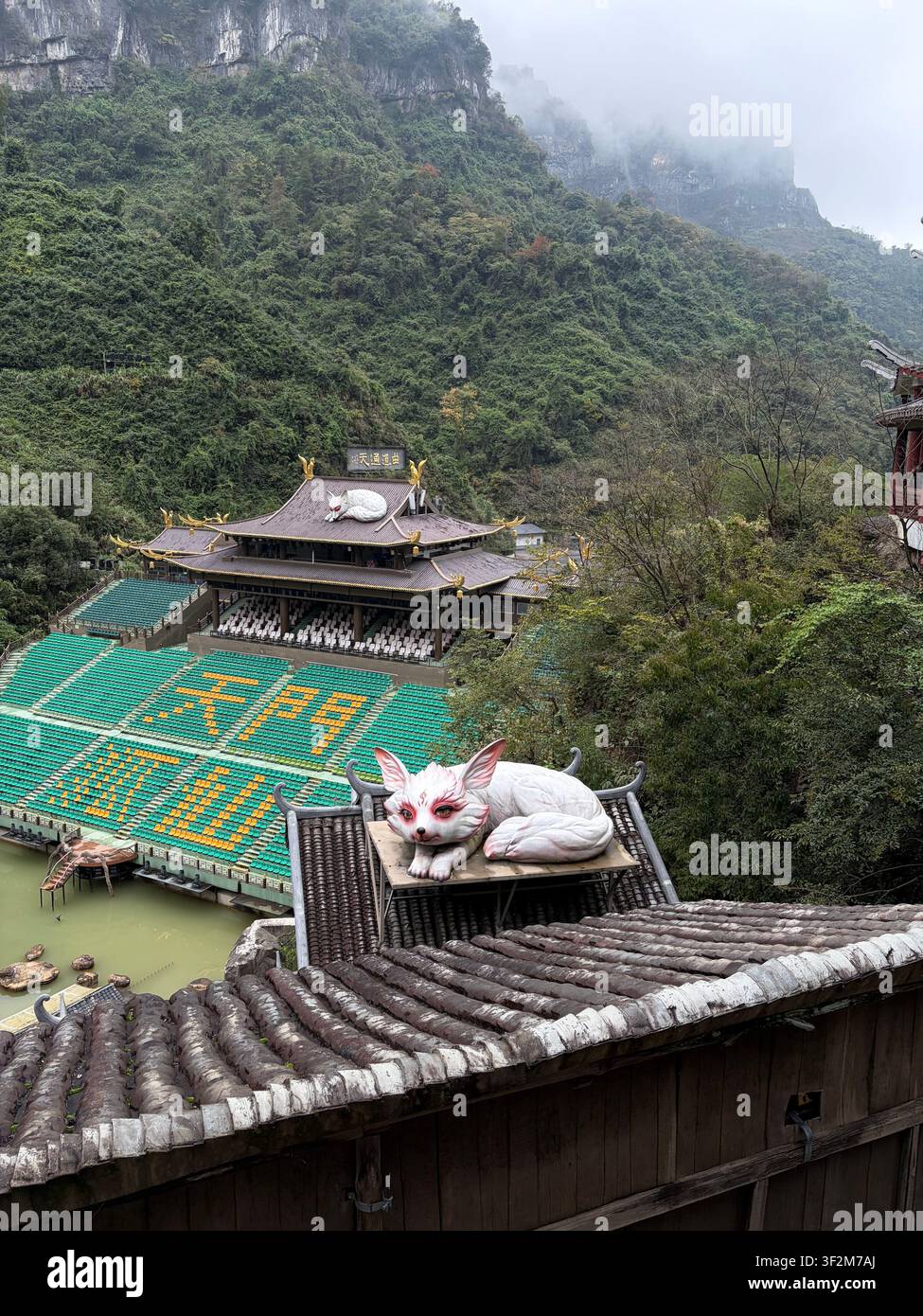 3 novembre 2025, Zhangjiajie, Chine – Sculpture mythologique d’un lapin blanc géant au sommet du toit d’une pagode chinoise traditionnelle avec des caractères jaunes, des falaises boisées brumeuses en arrière-plan. - Image de stock capturée avec un smartphone