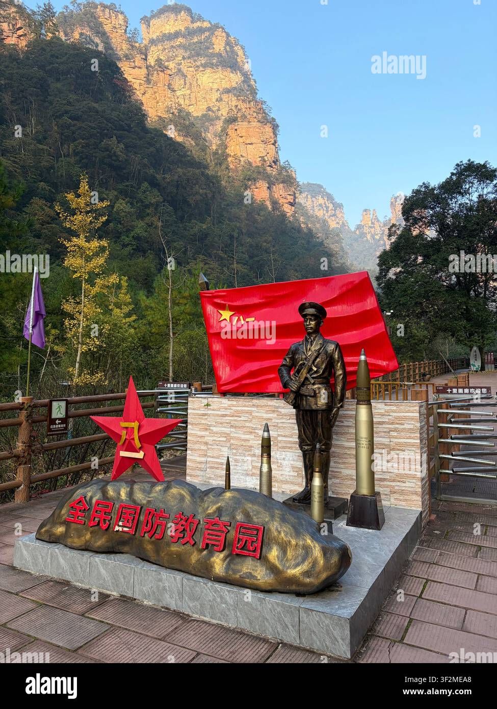 5 novembre 2025, Wulingyuan, Zhangjiajie, Chine – Statue en bronze d’un soldat avec drapeau rouge du PCC, obus d’artillerie et inscription patriotique chinoise devant des piliers karstiques en grès. Banque D'Images