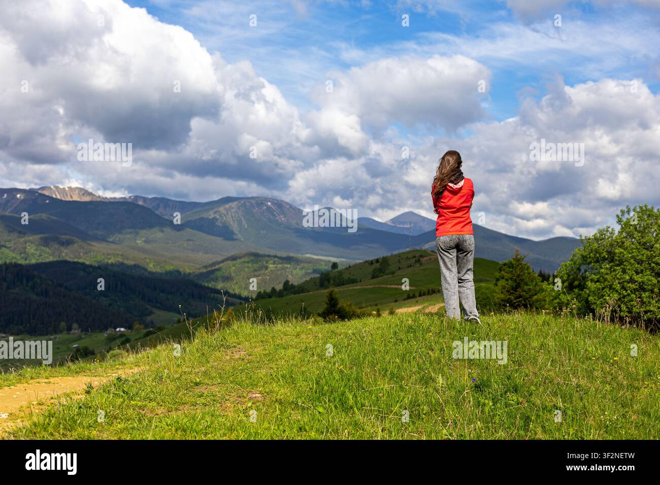 Femme surplombant une chaîne de montagnes depuis une colline herbeuse. Une femme en veste rouge se tient sur une colline herbeuse surplombant une vaste chaîne de montagnes en contrebas Banque D'Images