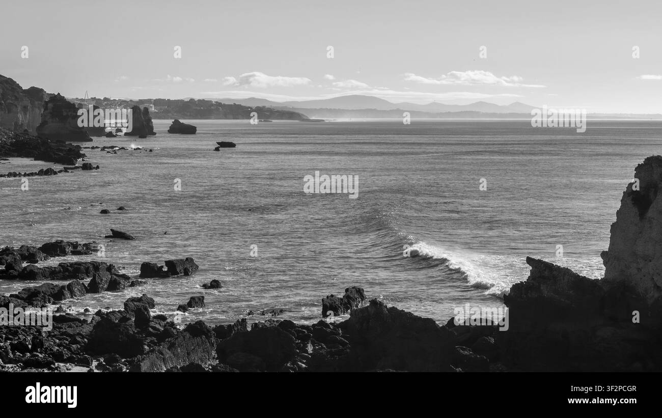 Les vagues s'écrasent contre les rochers le long de la côte. La scène capture un ciel clair et des collines lointaines en arrière-plan. L'heure de la journée semble être tôt Banque D'Images