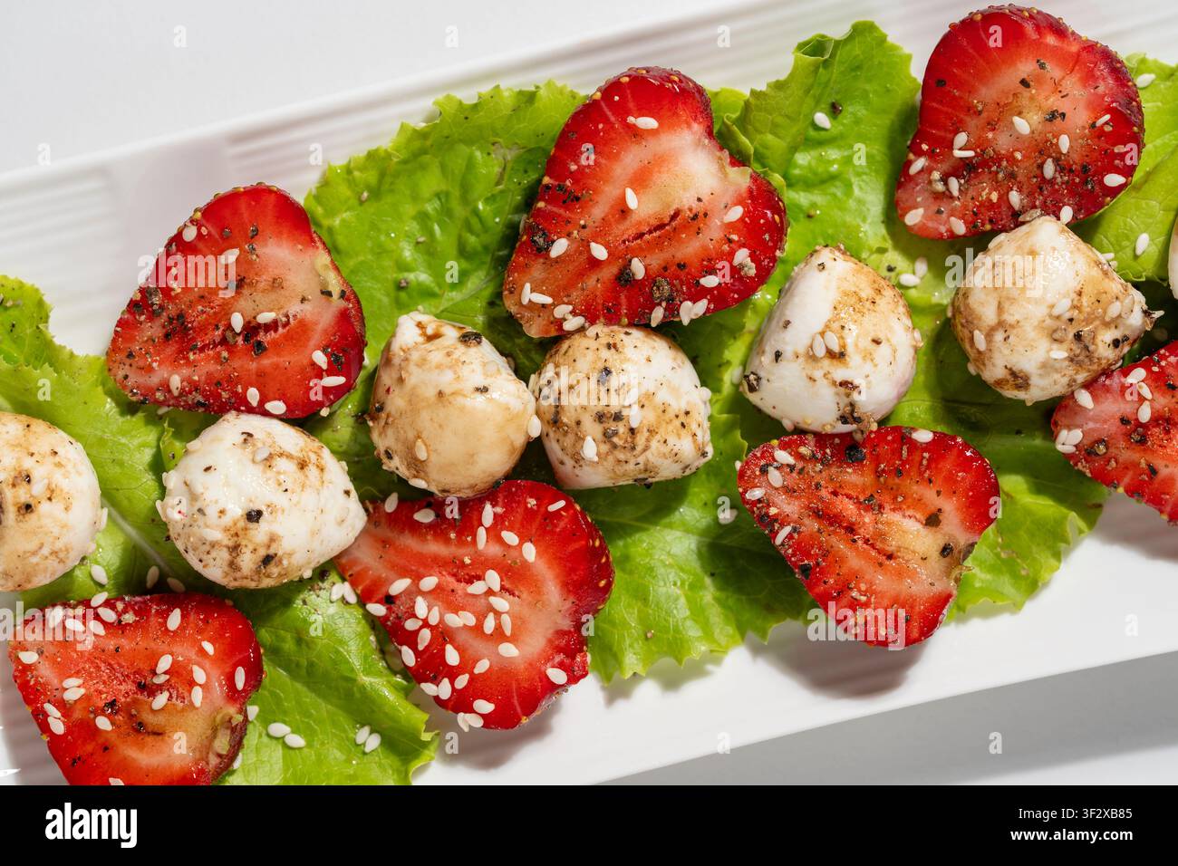 Salade d'été gourmande avec des fraises mûres, des boules de fromage mozzarella, de la laitue verte, une glacée au balsamique, du poivre noir et des graines de sésame sur une assiette blanche, gros plan Banque D'Images