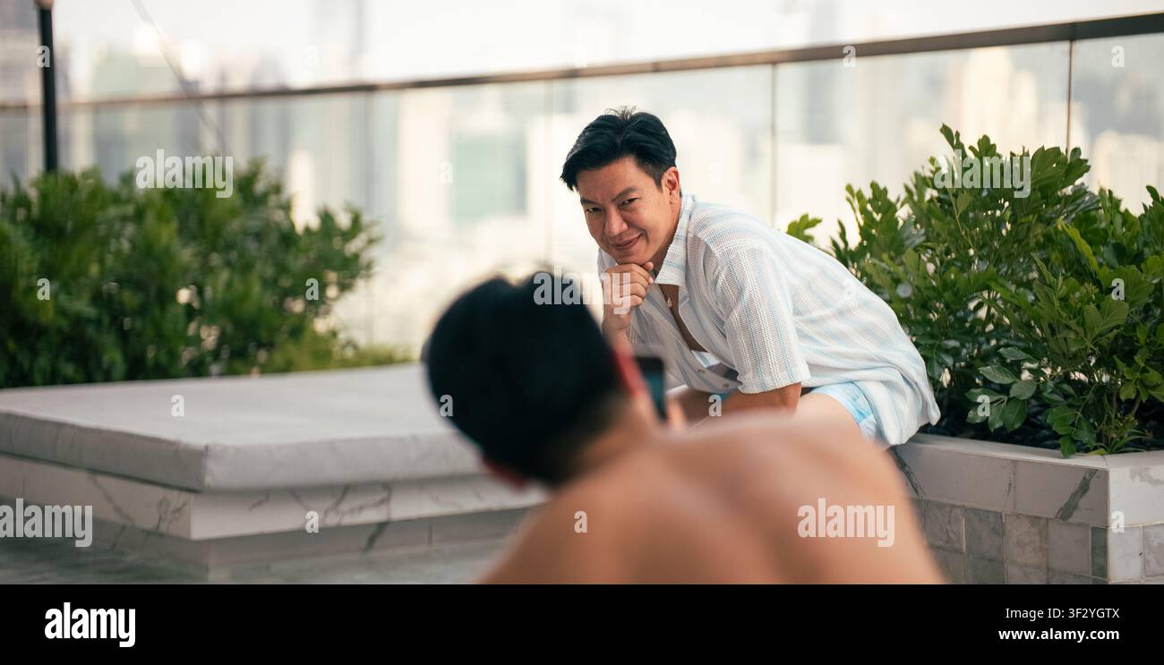 Homme asiatique souriant à son partenaire qui prend une photo, couple gay profitant de la piscine sur le toit Banque D'Images