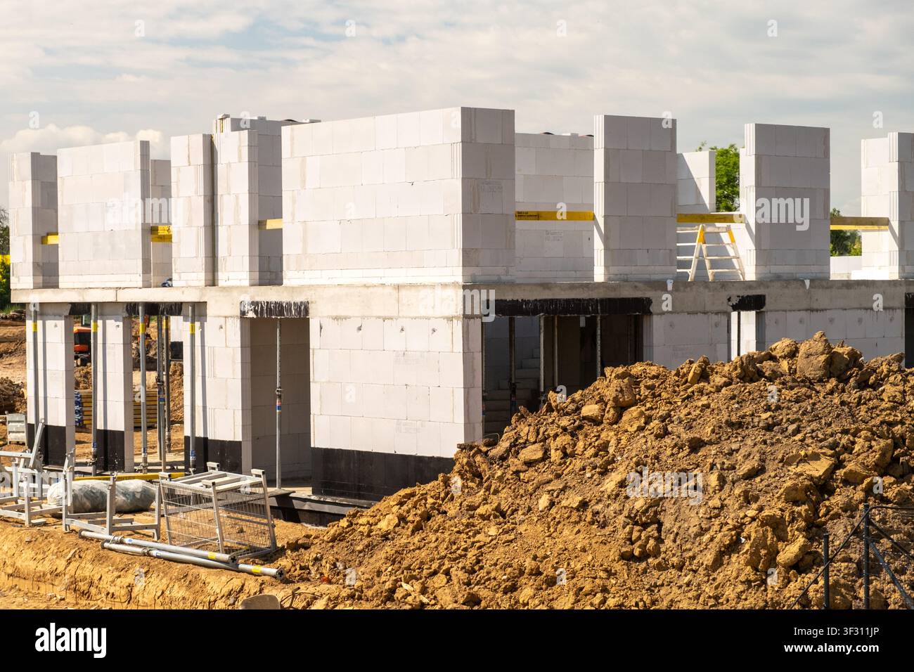 Processus de construction en cours avec des murs en blocs de béton en cours de montage sur le chantier. Des machines lourdes et des travaux de terrassement visibles au premier plan, indiquant un développement actif. Banque D'Images