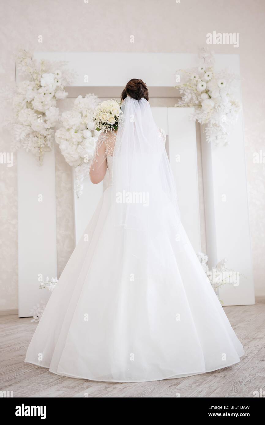 Mariee en robe de mariée blanche et voile tenant un bouquet devant une arche fleurie Banque D'Images