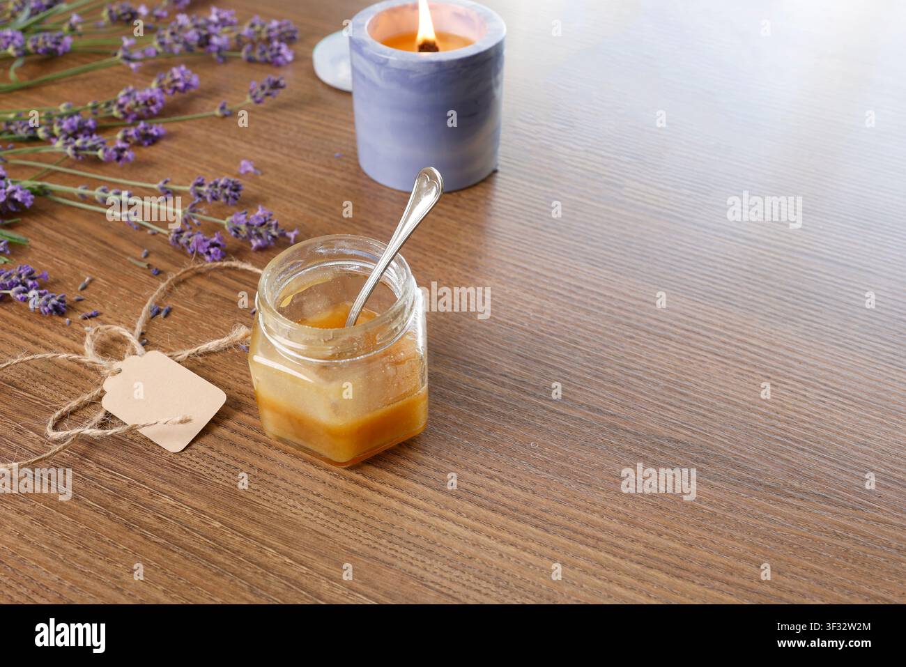 Bocal en verre de miel avec cuillère en métal sur fond de table en bois. Bougie parfumée brûlant près de tiges de bouquet de lavande fraîche. Composition de bien-être chaleureuse Banque D'Images