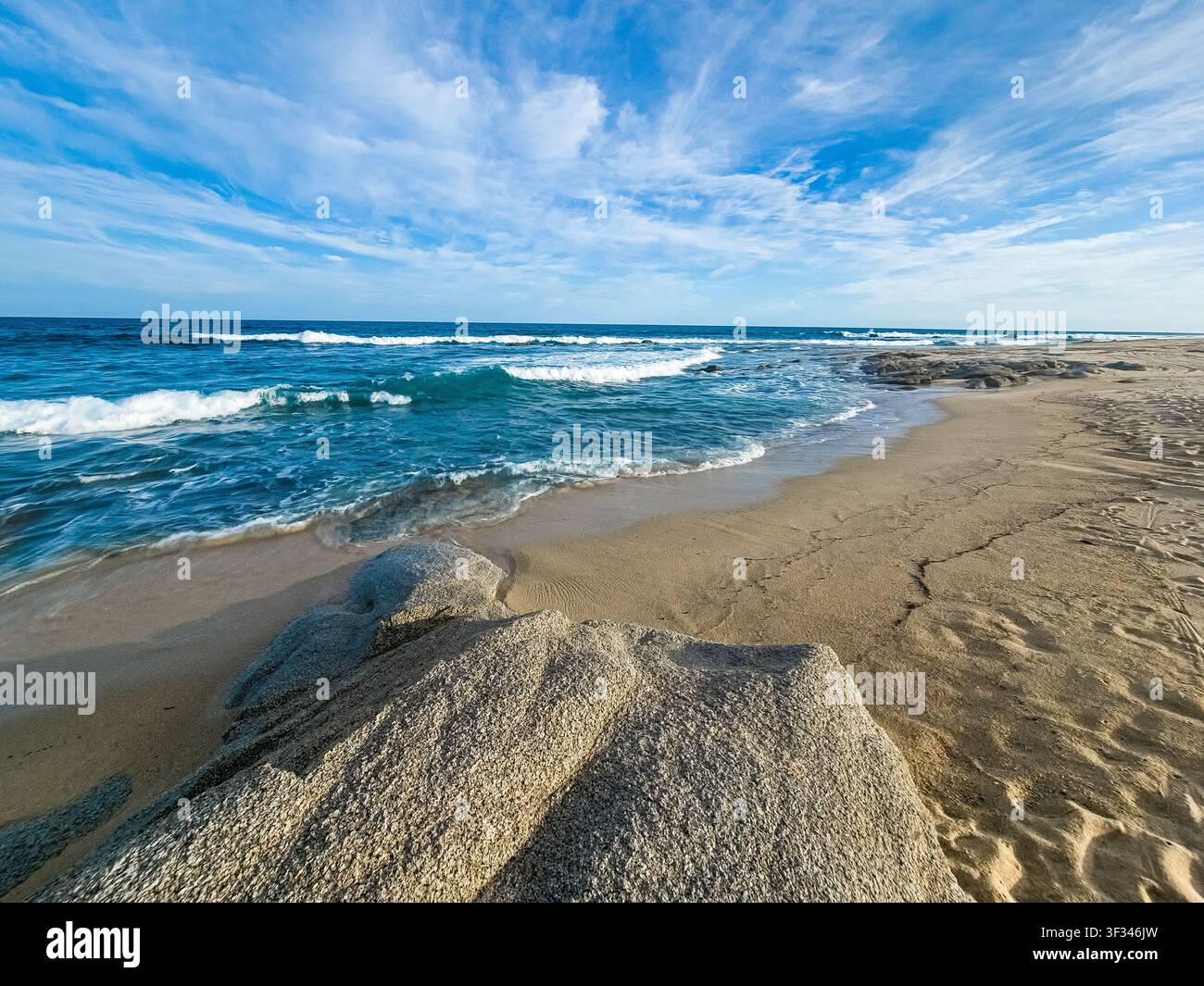 Sable doré rencontre des vagues turquoise sous un ciel bleu éclatant. Les vagues roulent doucement vers les rivages rocheux, laissant derrière elles des ondulations douces. Les nuages dérivent paresseusement Banque D'Images
