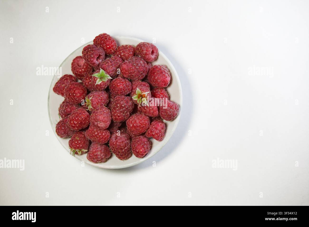 Une photographie en gros plan vu de dessus d'un bol en céramique blanc rond rempli de framboises rouges fraîches et mûres. Le bol est positionné sur le côté gauche d'une claire Banque D'Images