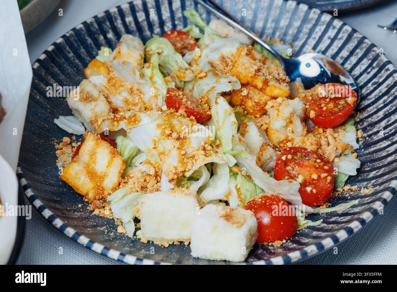 Bol rayé sombre rempli de salade. Chou râpé et tomates cerises se mélangent avec des croutons grillés. Des morceaux de protéines grillées ajoutent de la texture. Cuillère en métal r Banque D'Images