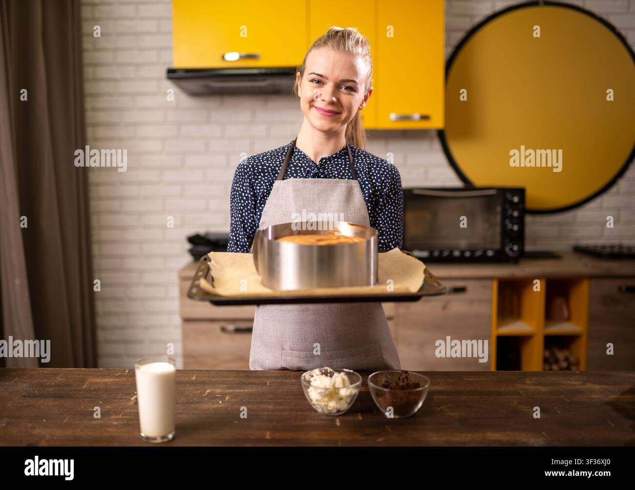Femme souriante tenant un gâteau rond fraîchement cuit sur un plateau dans une cuisine moderne aux armoires jaunes Banque D'Images