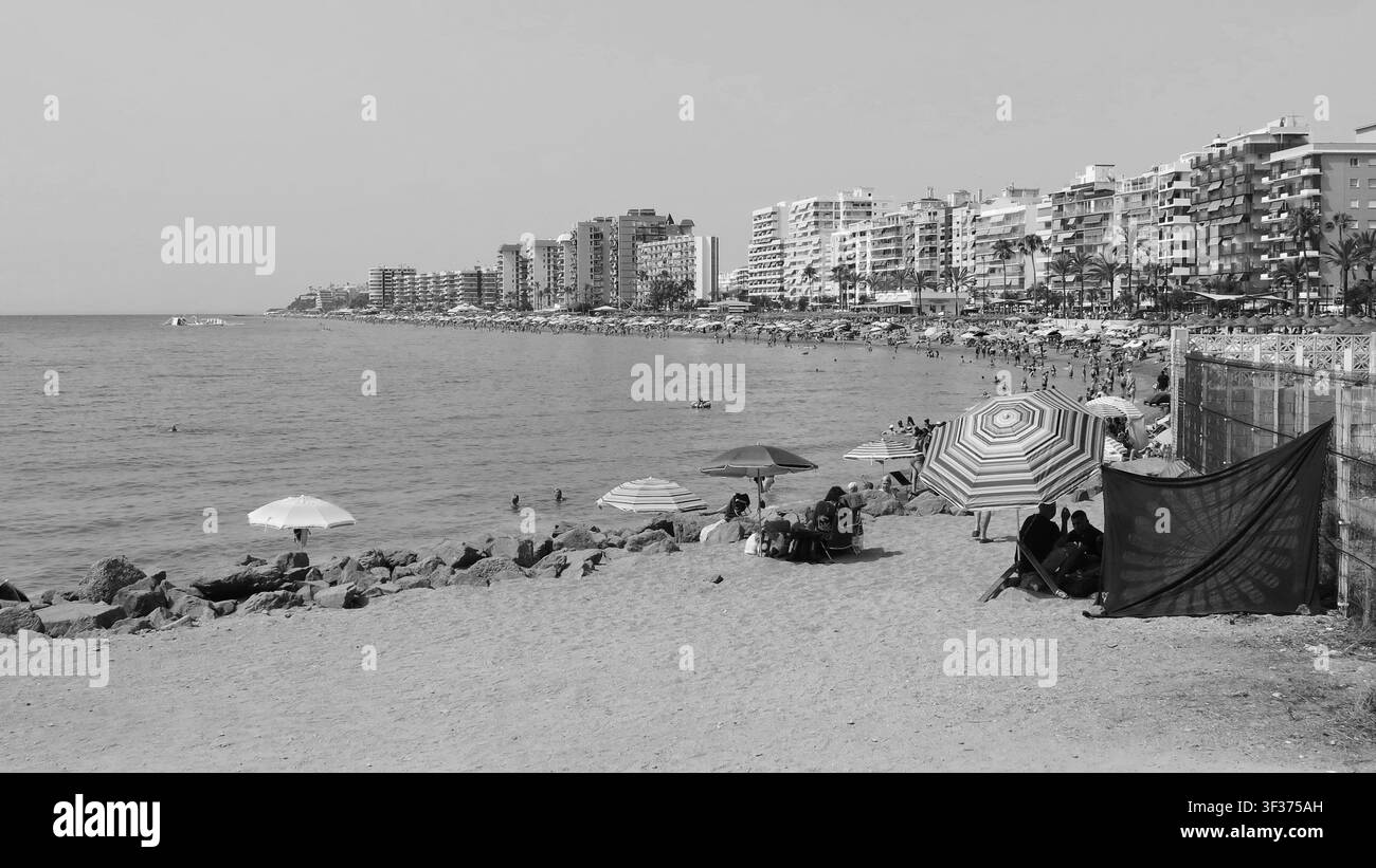 Vue en noir et blanc d'une plage de sable bondée à Fuengirola avec des parasols, des touristes et des appartements en hauteur le long de la côte de la Costa del Sol. Banque D'Images