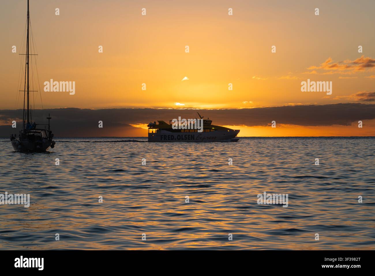 Une vedette rapide rouge et jaune de Fred Olsen Express traverse une mer dorée au coucher du soleil devant un yacht à voile en silhouette au large de La Gomera, Îles Canaries, Espagne, photographiée le 20 janvier 2026. Banque D'Images