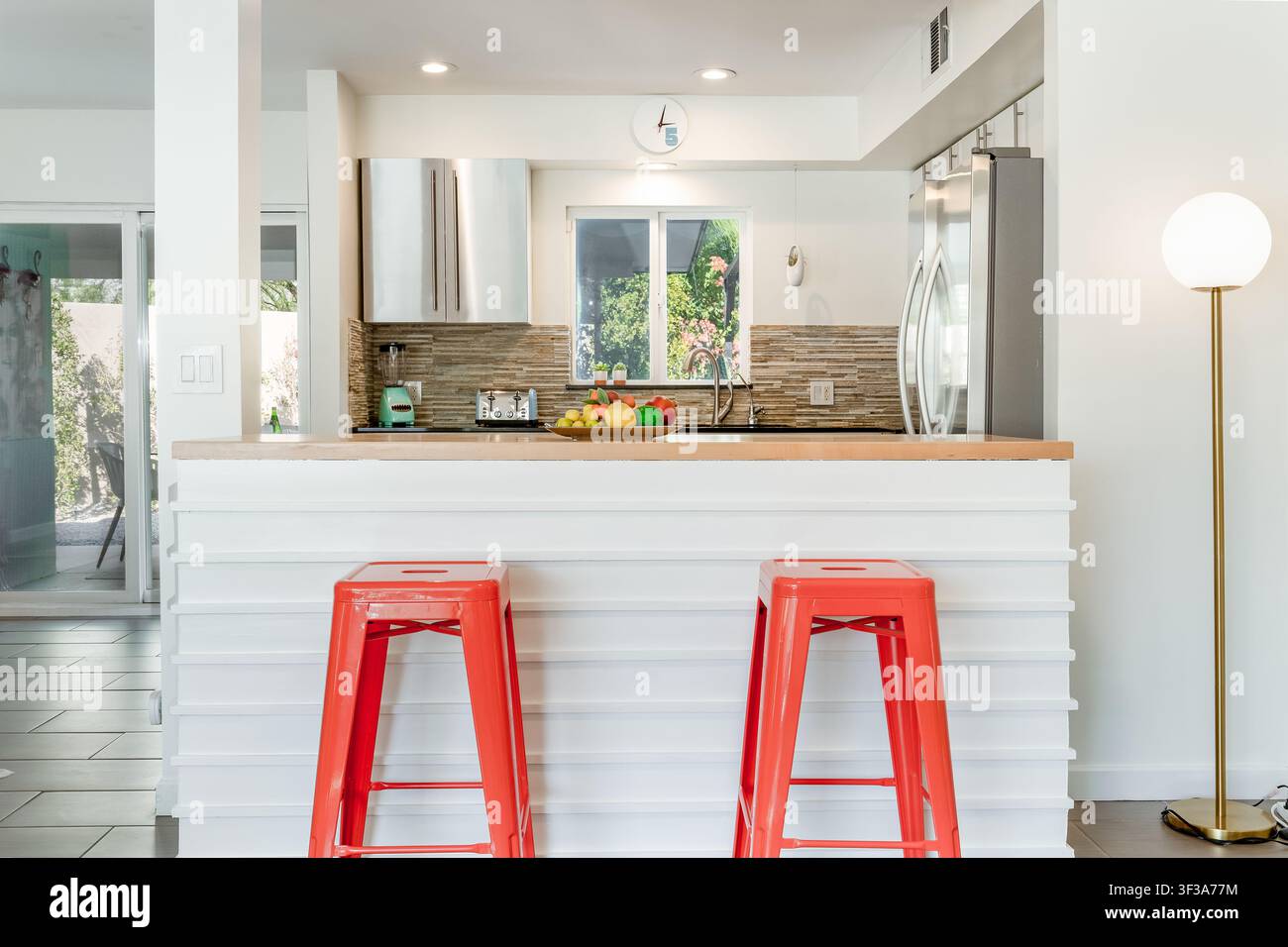 Une prise d'intérieur lumineuse et aérée d'une cuisine blanche moderne. La scène présente une île de comptoir blanc avec des tabourets rouges et un dosseret carrelé coloré Banque D'Images