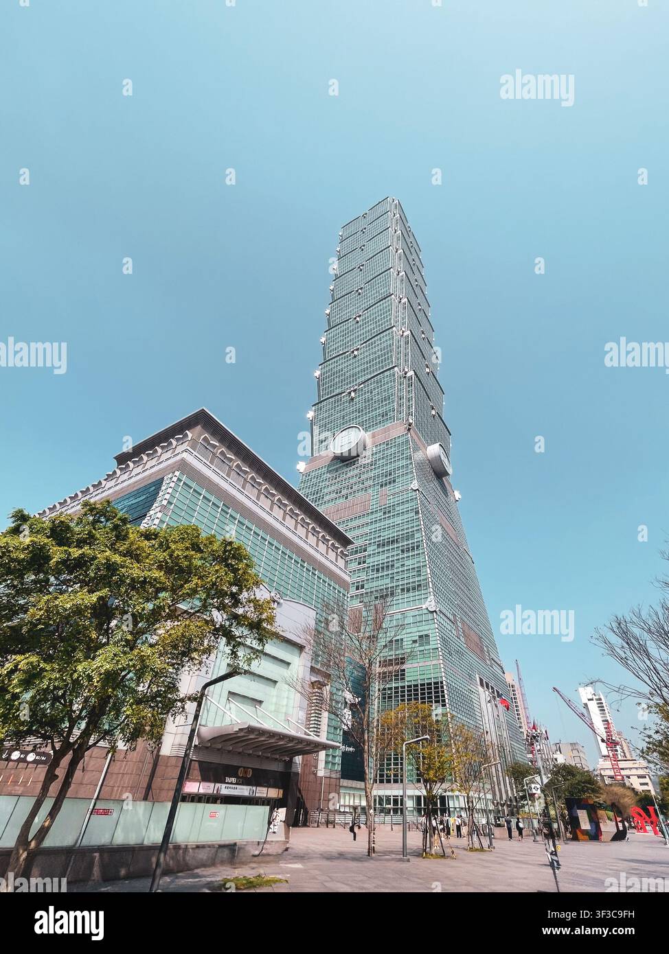 Taipei 101 s'élève de façon spectaculaire contre un ciel bleu clair dans une vue en contre-plongée depuis le district de Xinyi, Taipei. La tour emblématique à étages en verre vert domine l'horizon au-dessus des arbres et des blocs urbains environnants. Banque D'Images