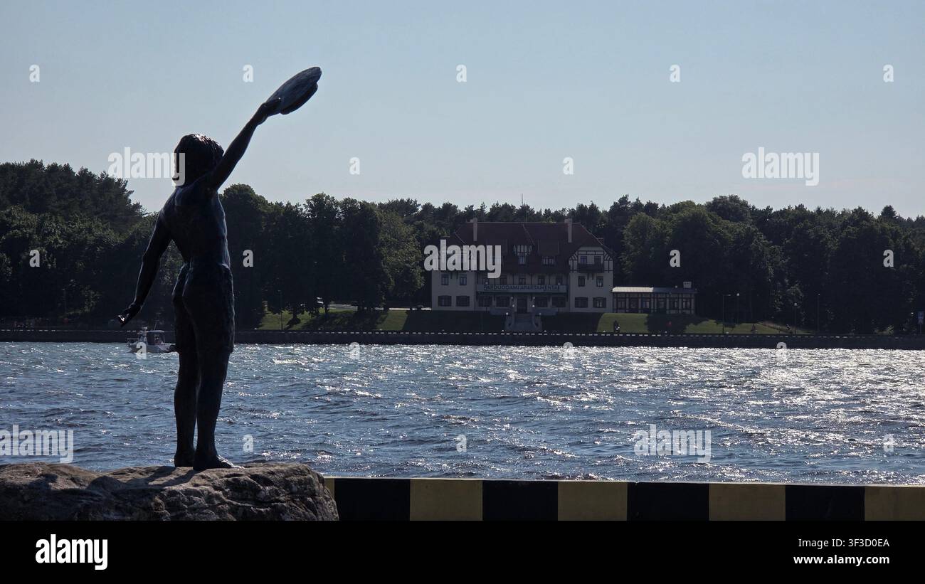 Silhouette de la statue « Vaikystės svajonė » sur l'embarcadère de Klaipėda avec la lagune curonne étincelante et une villa historique sur la presqu'île de Curonie, Lituanie. Banque D'Images