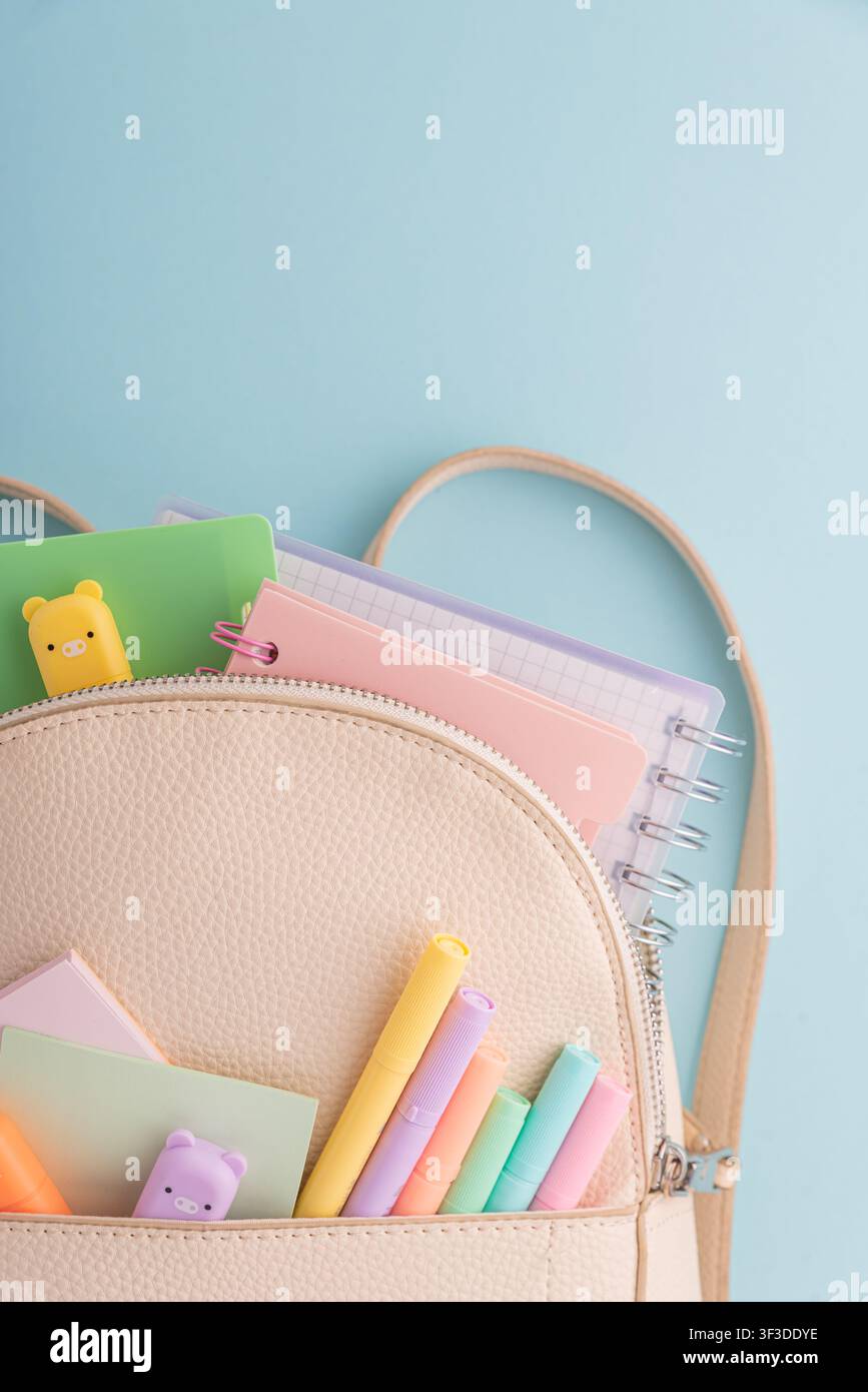 Ouverture d'un sac à dos révélant une collection de cahiers aux couleurs pastel, des stylos, des surligneurs, des notes adhésives et des gommes, symbolisant l'éducation, la créativité, Banque D'Images