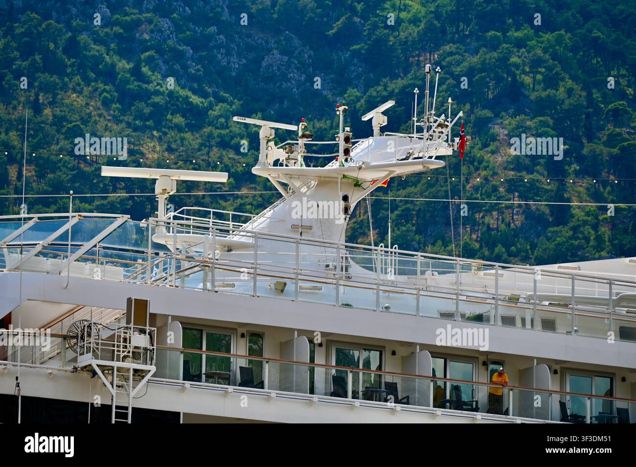 Prise de vue détaillée de la tour radar et du mât de navigation d'un croisière blanc moderne, équipé de dispositifs de communication, de drapeaux de signalisation et de balcons supérieurs des passagers, surplombant une colline montagneuse luxuriante. Banque D'Images