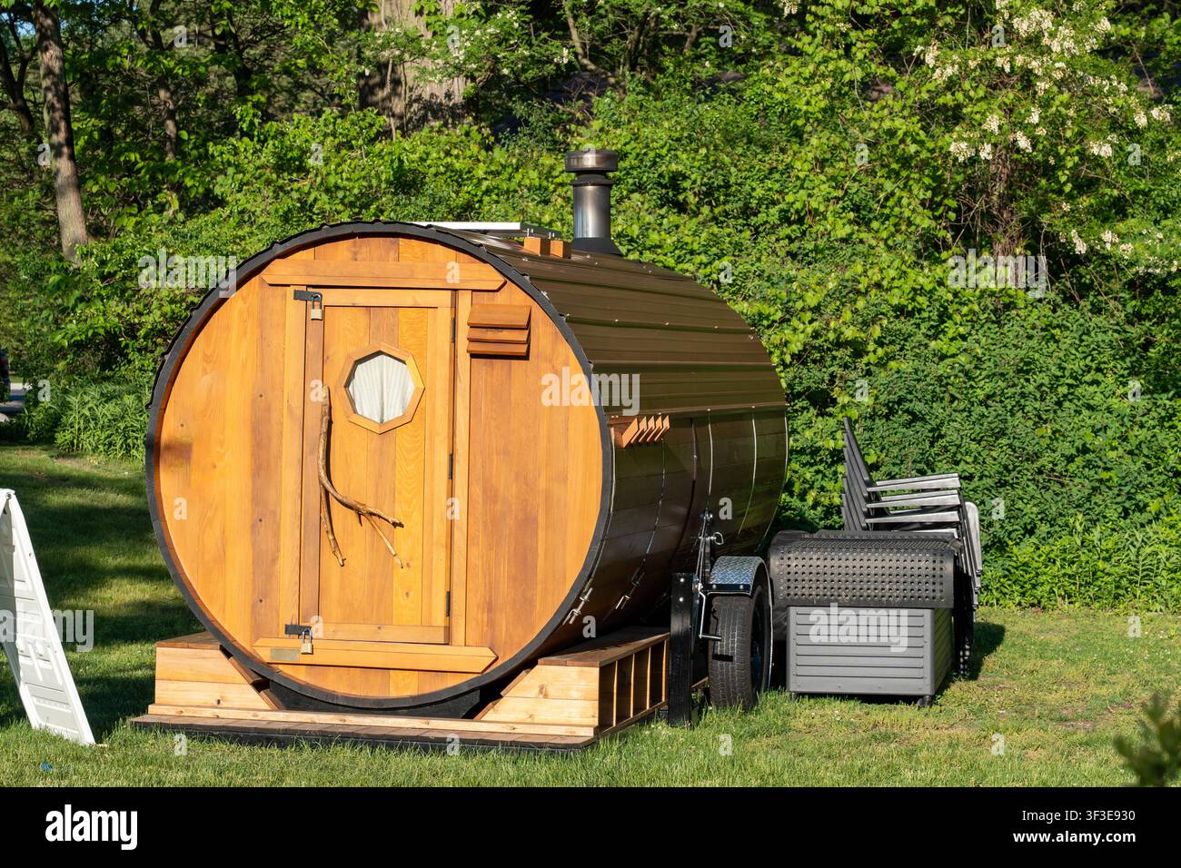Sauna portable en forme de tonneau chauffée au bois sur une remorque entourée d'une végétation luxuriante, avec une façade en bois, une cheminée et une zone de détente extérieure Banque D'Images