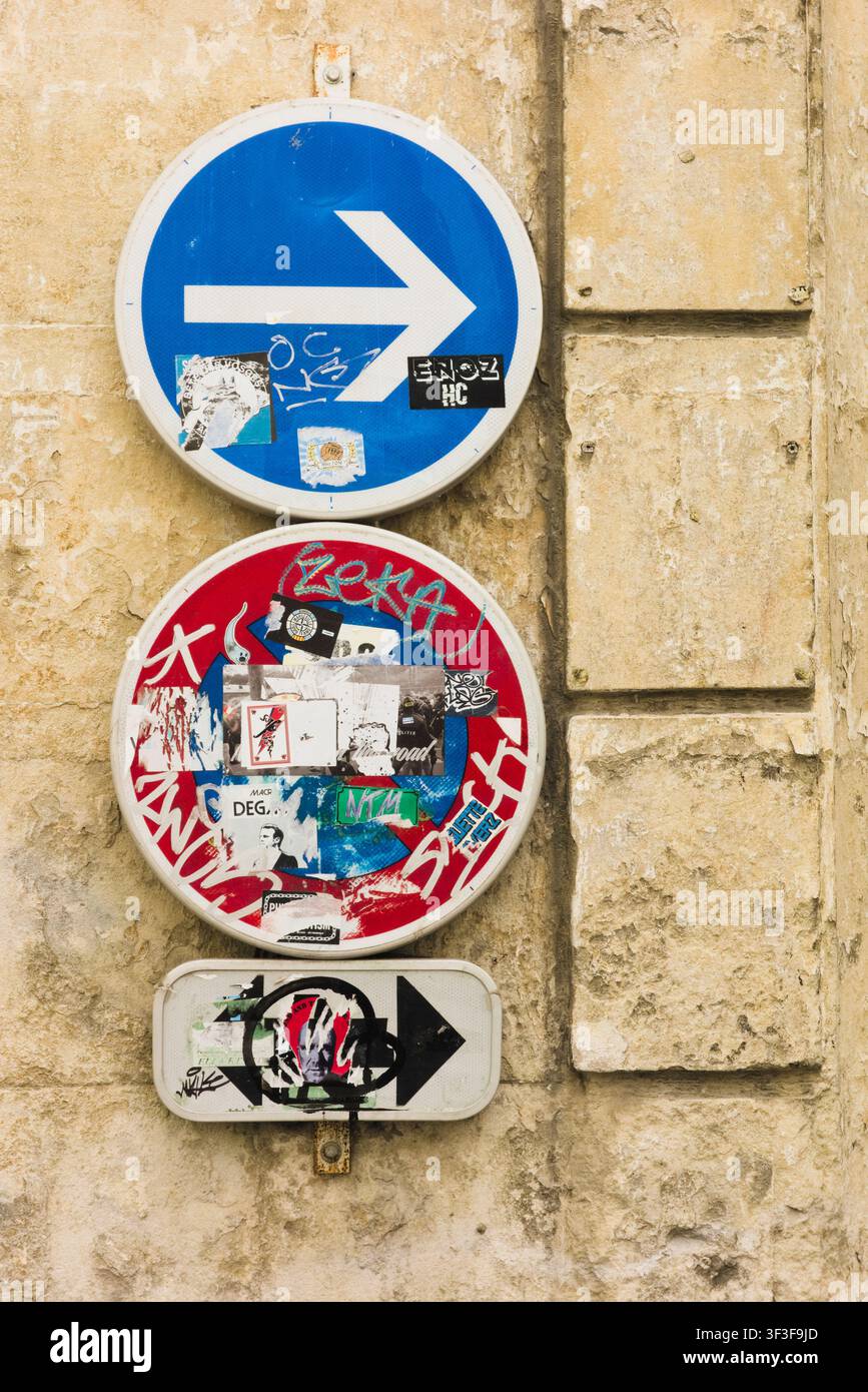 Close-up de panneaux de signalisation routière circulaires recouverts d'autocollants urbains et de graffitis montés sur un mur de pierre rustique à Arles, France. Banque D'Images