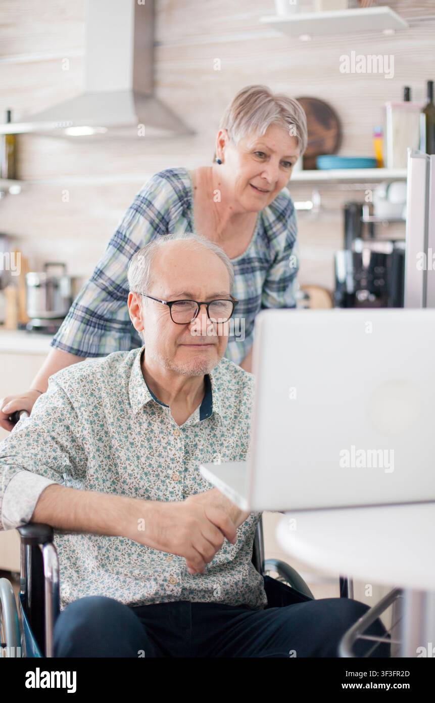 Un homme âgé handicapé en fauteuil roulant passe un appel vidéo familial avec sa femme sur un ordinateur portable dans la cuisine, utilisant la communication en ligne à domicile. Banque D'Images