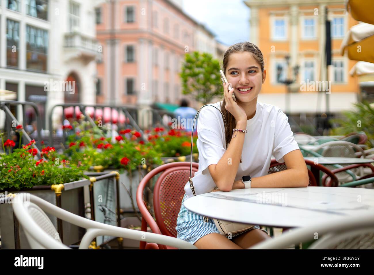 Jeune femme souriante téléphonant sur smartphone dans un café en plein air. Jeune femme souriante parle au smartphone tout en étant assise dans un café en plein air entourée de Banque D'Images