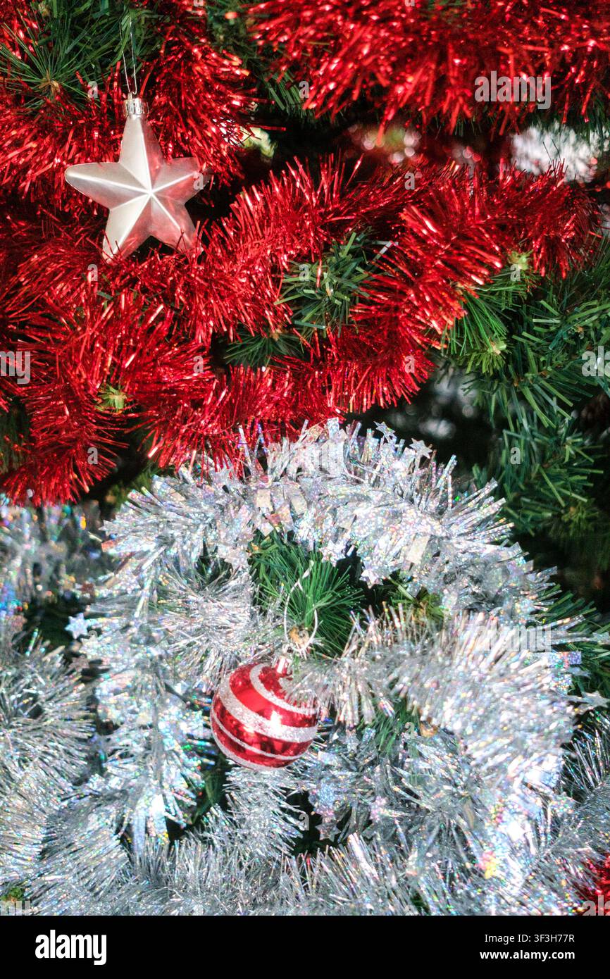 Vue rapprochée de décorations de Noël, de guirlandes rouges, de guirlande argentée, d'un ornement en forme d'étoile et d'un objet artisanal de Noël en papier posé sur un arbre dans une maison familiale à Banque D'Images