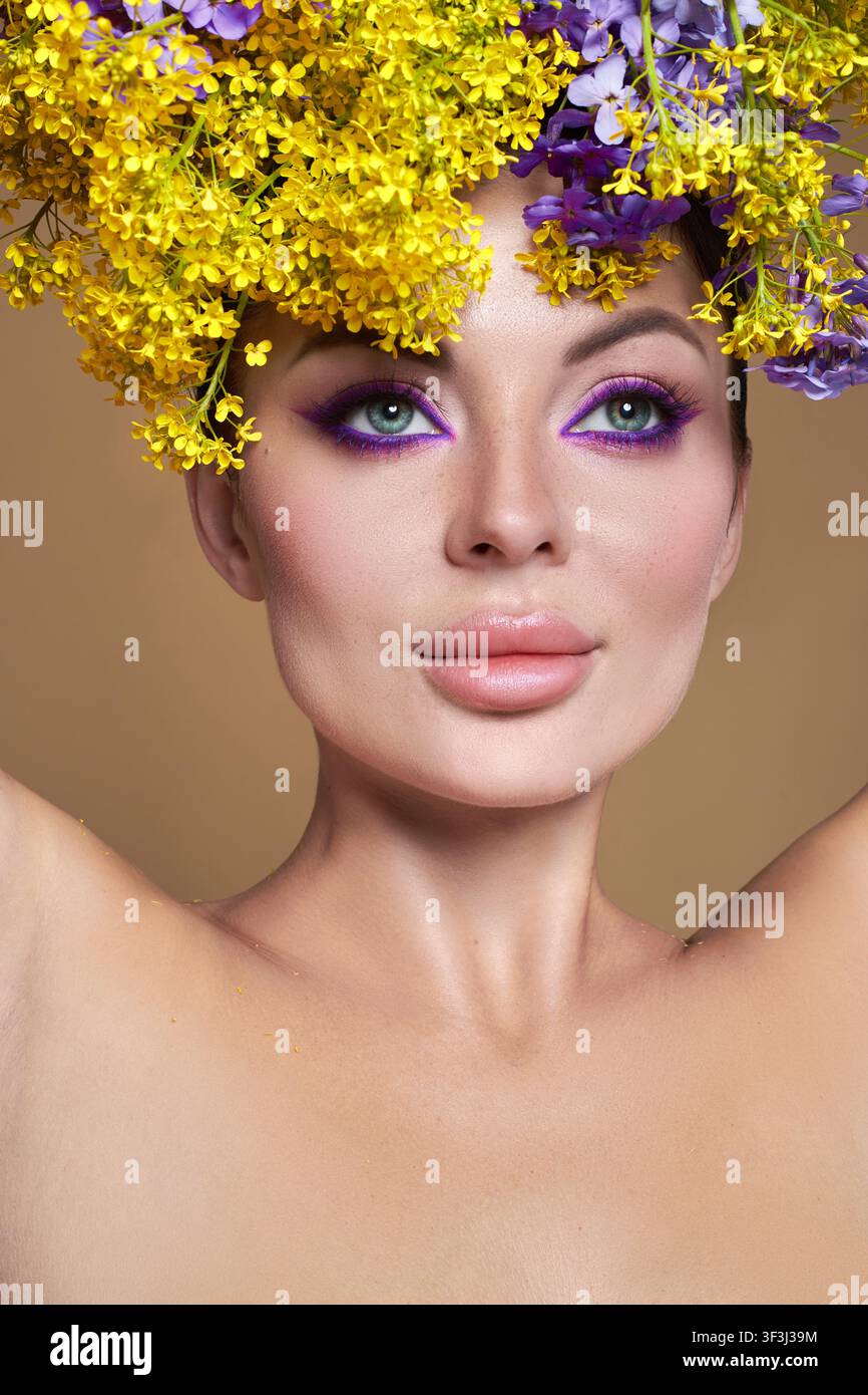 Modèle de beauté debout devant un fond simple. Elle porte une couronne faite de fleurs jaune vif et violettes. Son maquillage présente des couleurs audacieuses qui com Banque D'Images