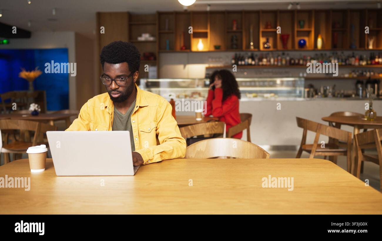 Homme afro-américain concentré portant des lunettes travaillant sur un ordinateur portable dans un café et buvant une boisson chaude. Homme assis à une table en bois avec une tasse de café, femme Banque D'Images