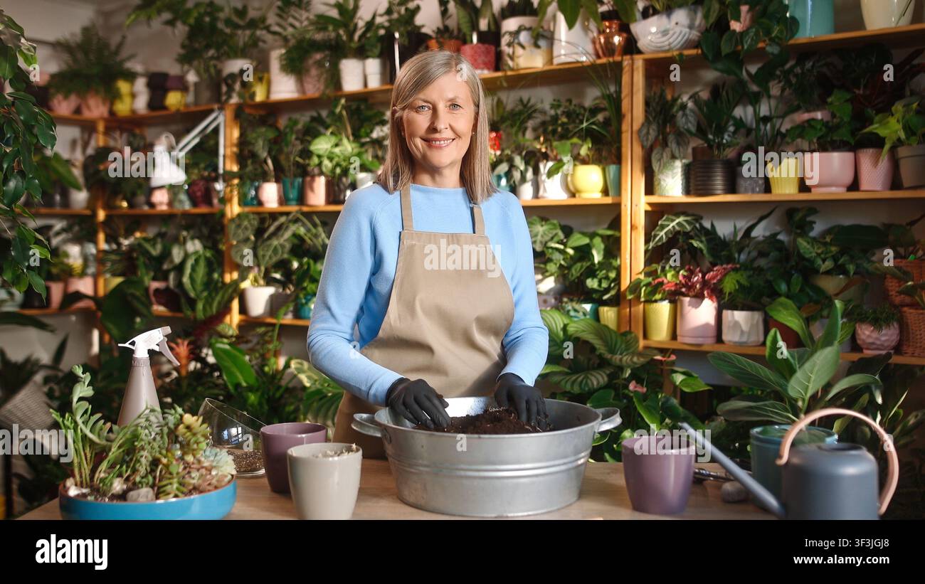 Portrait d'une belle femme mûre attentionnée travaillant dans une fleuristerie et prenant soin des plantes et de la terre. Attrayante femme âgée aux cheveux gris heureuse caucasienne Banque D'Images