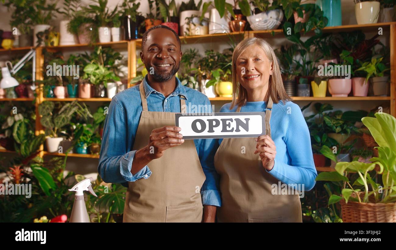 Portrait de deux fleuristes joyeux et prospères regardant l'appareil photo en souriant, tenant un panneau « Ouvert », portant un tablier et appréciant leur travail à la boutique de fleurs. Jeune Africain Banque D'Images