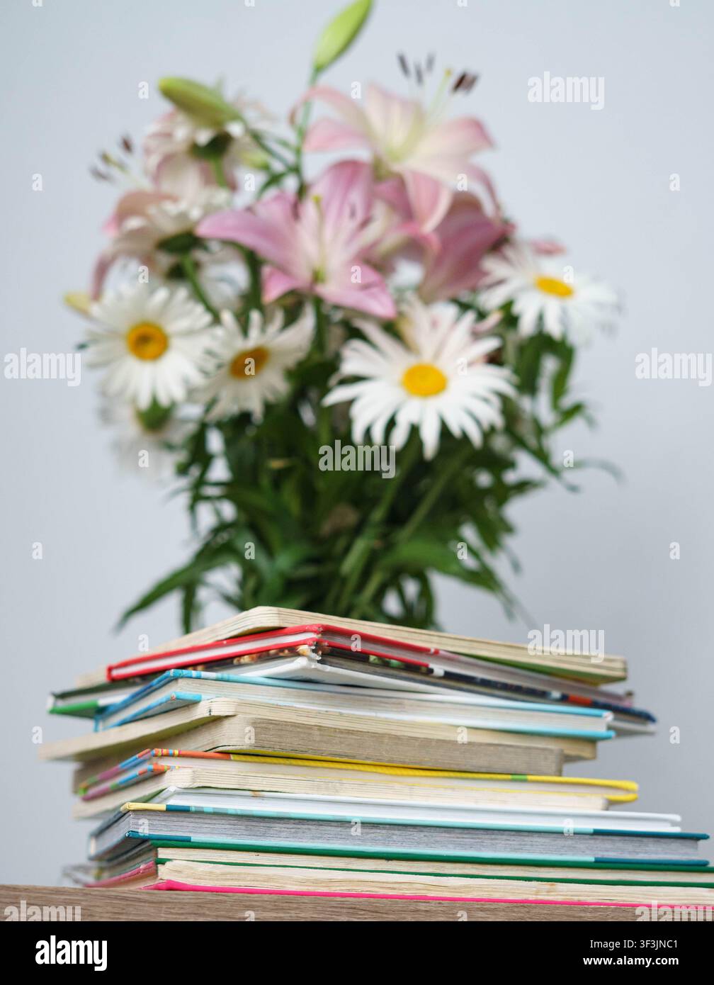 Pile de livres colorés sur une table en bois avec un bouquet de lys et de marguerites en fleurs en arrière-plan. Banque D'Images