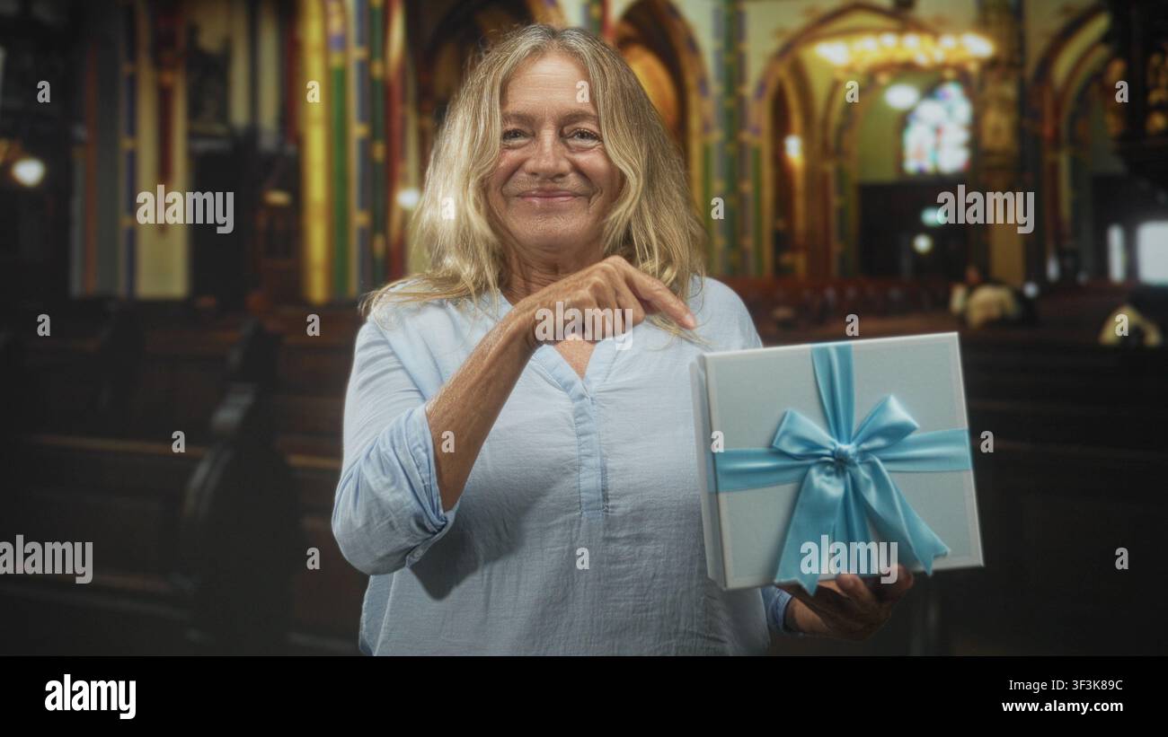 Femme senior blonde pointe le doigt à la boîte cadeau emballée avec la main visible tout en souriant et tenant présent à l'intérieur du bâtiment orné ; contentement chaleureux. Banque D'Images