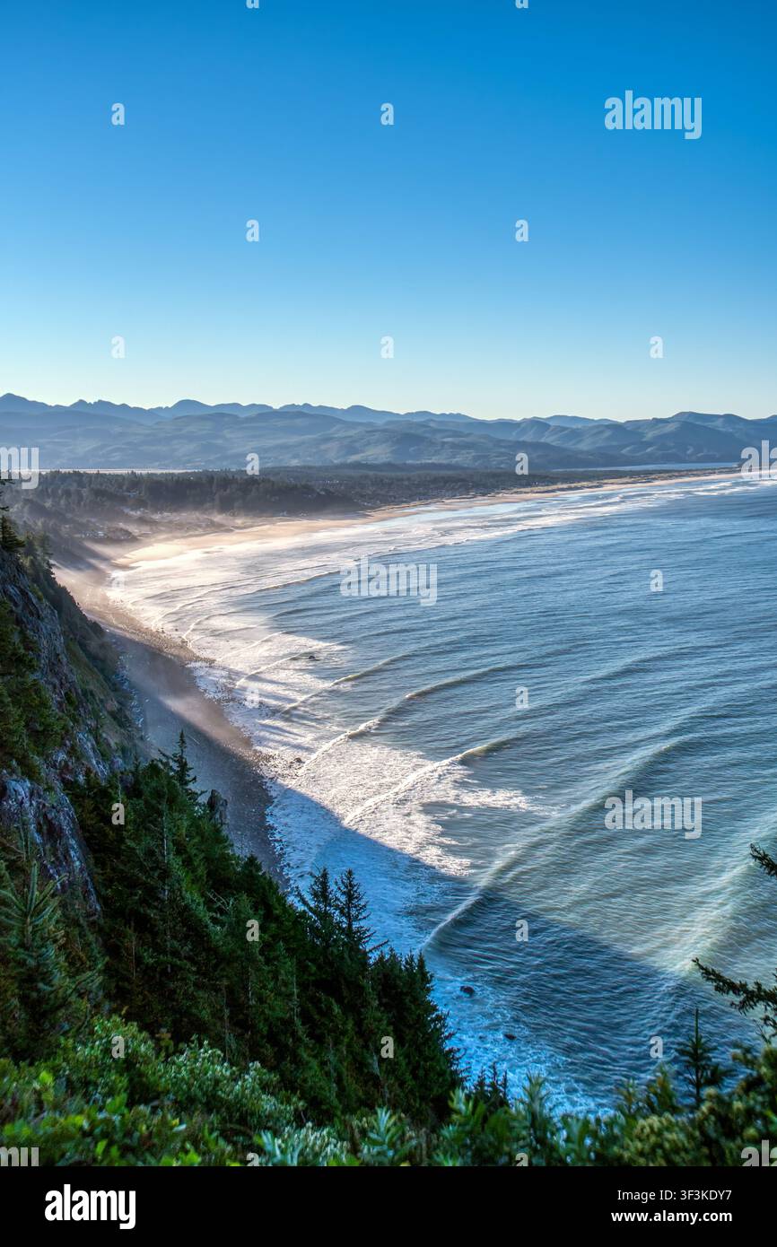 Un ciel bleu vif rencontre une côte accidentée. Les vagues se brisent doucement contre le rivage sablonneux. Les falaises s'élèvent abruptement, drapées d'une végétation dense. La brume tournoie près de l'eau Banque D'Images