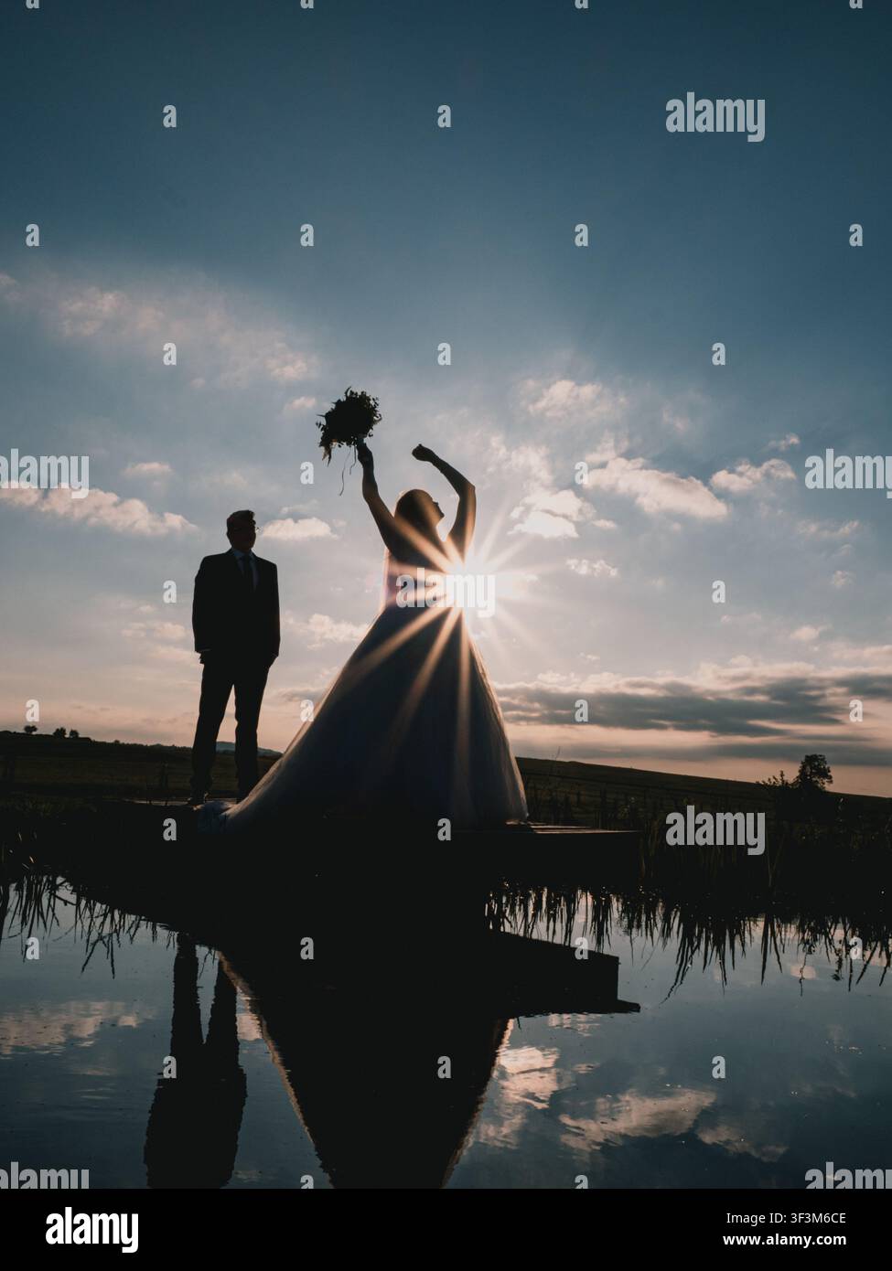 Silhouettes de mariage avec éclat de soleil et reflets sur une jetée en bois au-dessus de l'eau calme au coucher du soleil. Banque D'Images