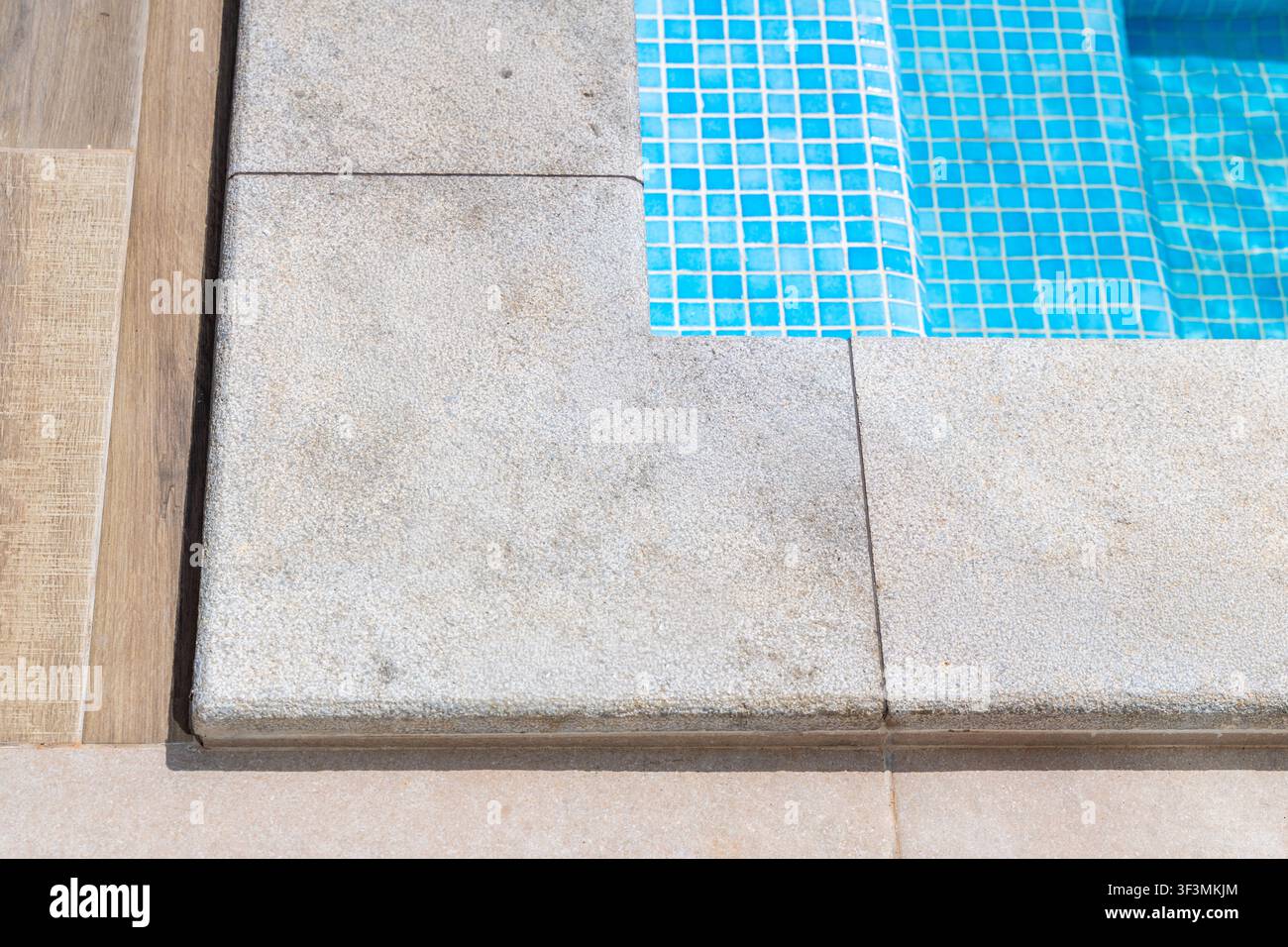 Vue détaillée de la transition entre une terrasse en bois et un rebord de piscine en granit ainsi que des marches carrelées en bleu menant à une eau claire Banque D'Images