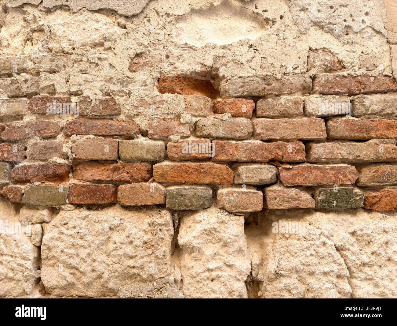 Un gros plan d'un mur en plâtre vieilli révèle une tache de brique rouge exposée parmi des pierres naturelles rugueuses, usées par des décennies de temps. Banque D'Images