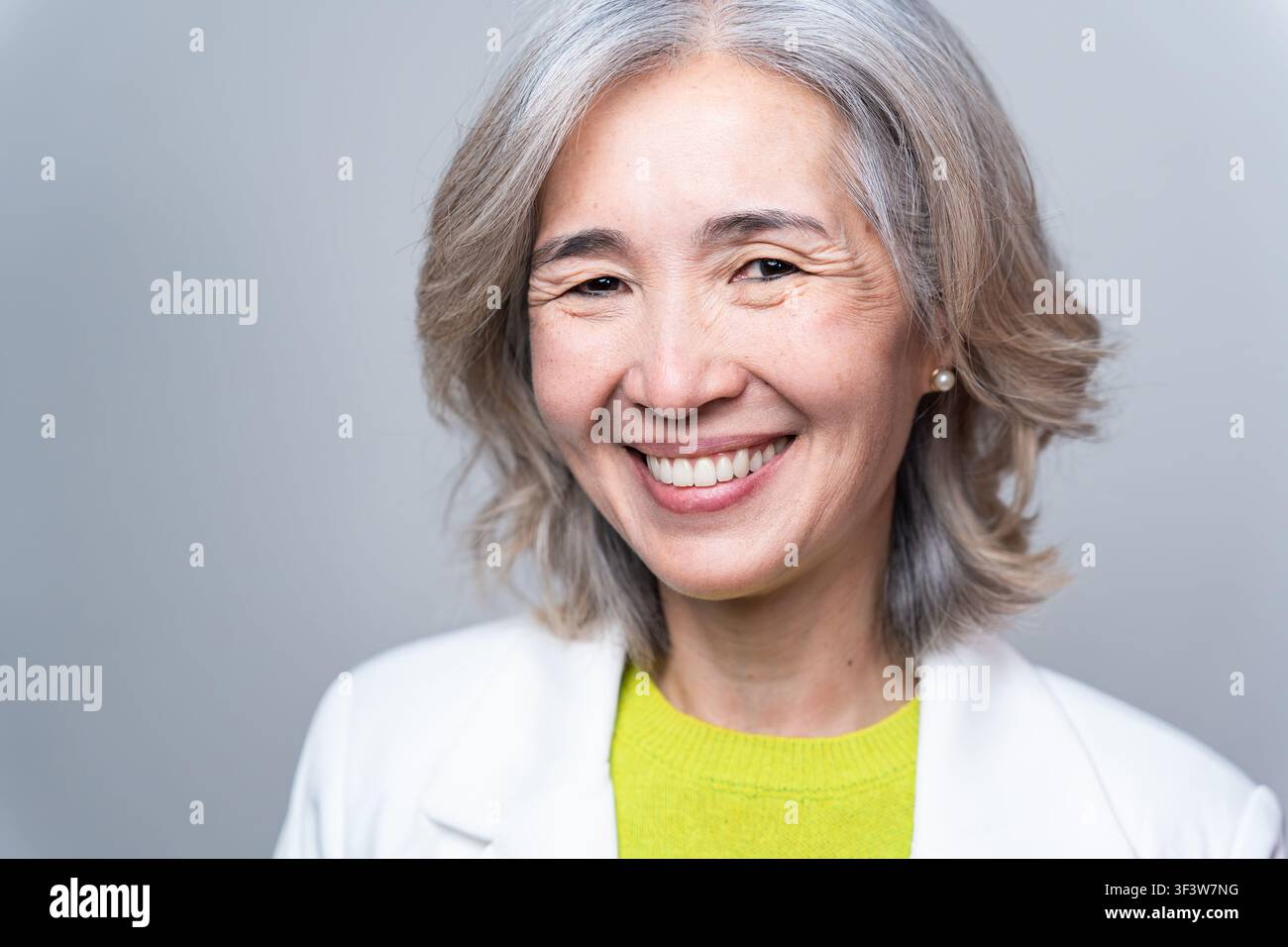 Portrait studio chaleureux d'une femme asiatique âgée souriante aux cheveux gris, portant un blazer blanc et un haut vert, regardant confiante et amicale contre un si Banque D'Images