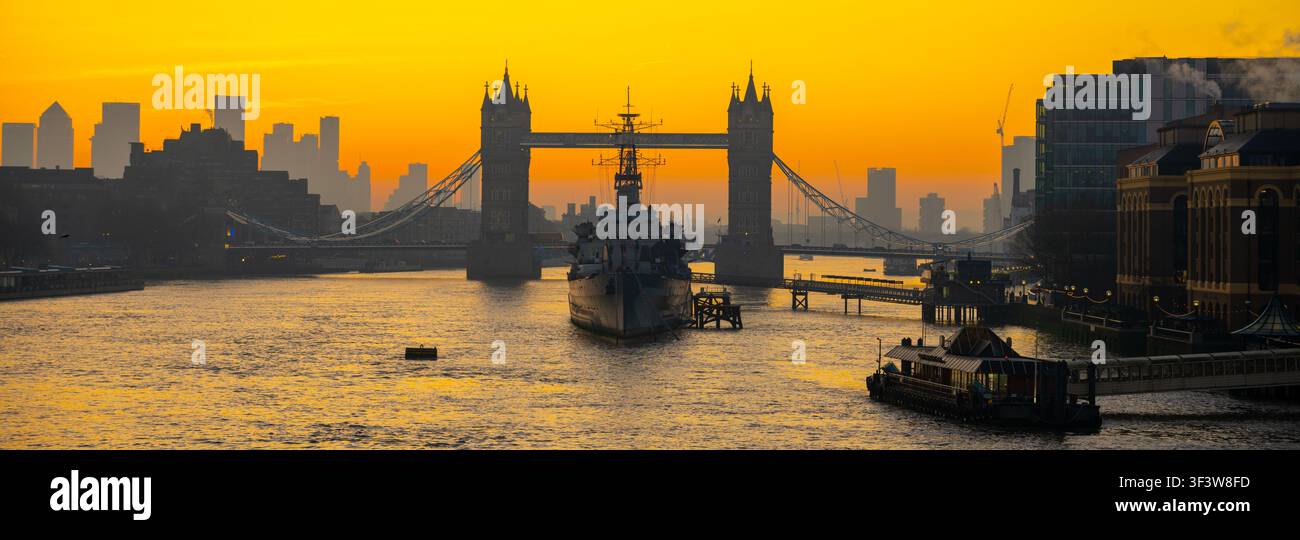 La lumière matinale éclaire la Tamise. Le HMS Belfast est ancré sur l'eau tandis que le Tower Bridge se dresse en silhouette. La ligne d'horizon de la ville dessine la scène, créant une vue visuelle dynamique. Banque D'Images