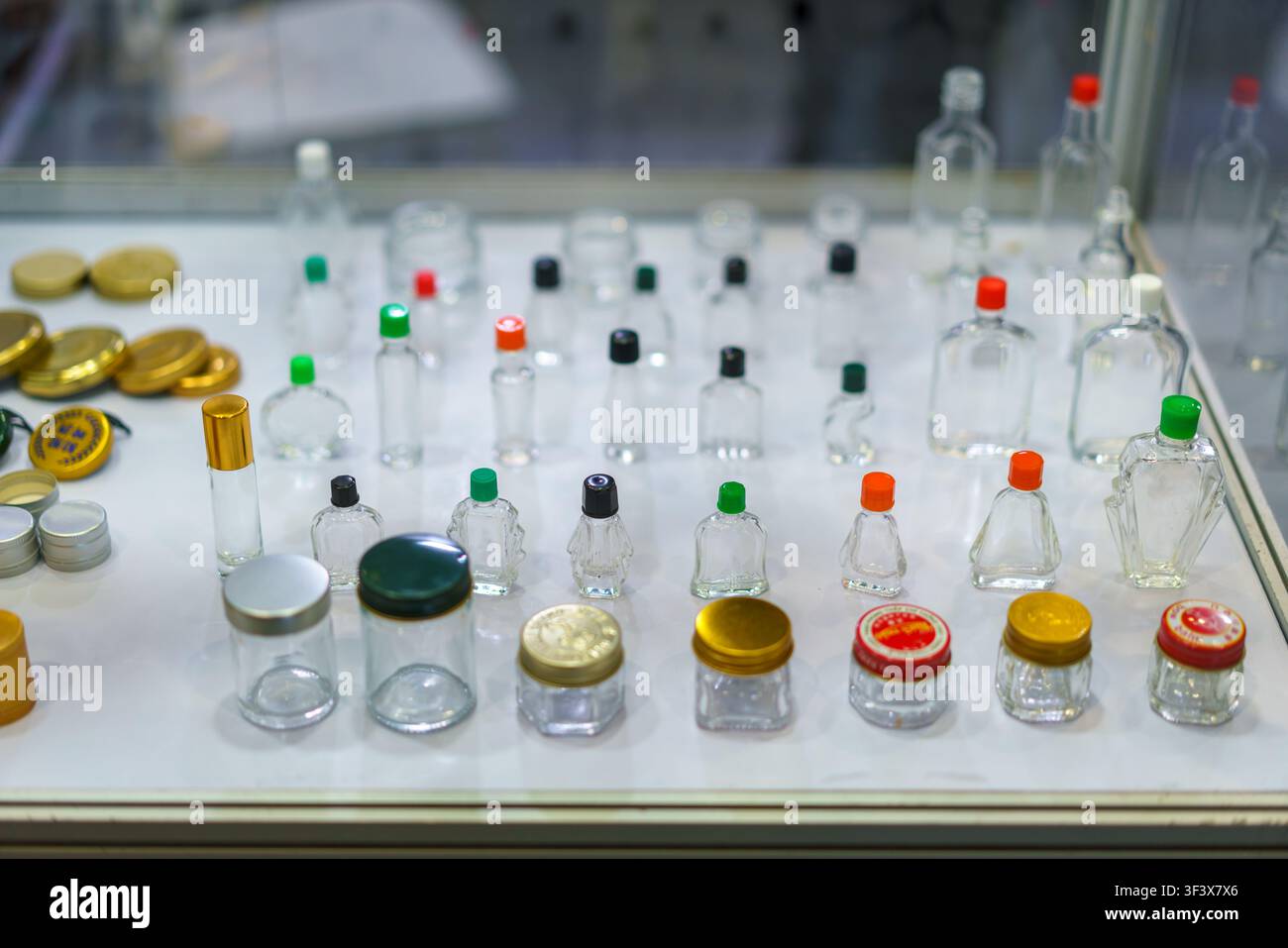 Une collection variée de petites bouteilles en verre vides et de pots de cosmétiques avec des bouchons colorés disposés sur une table d'exposition blanche, mettant en valeur différents emballages Banque D'Images