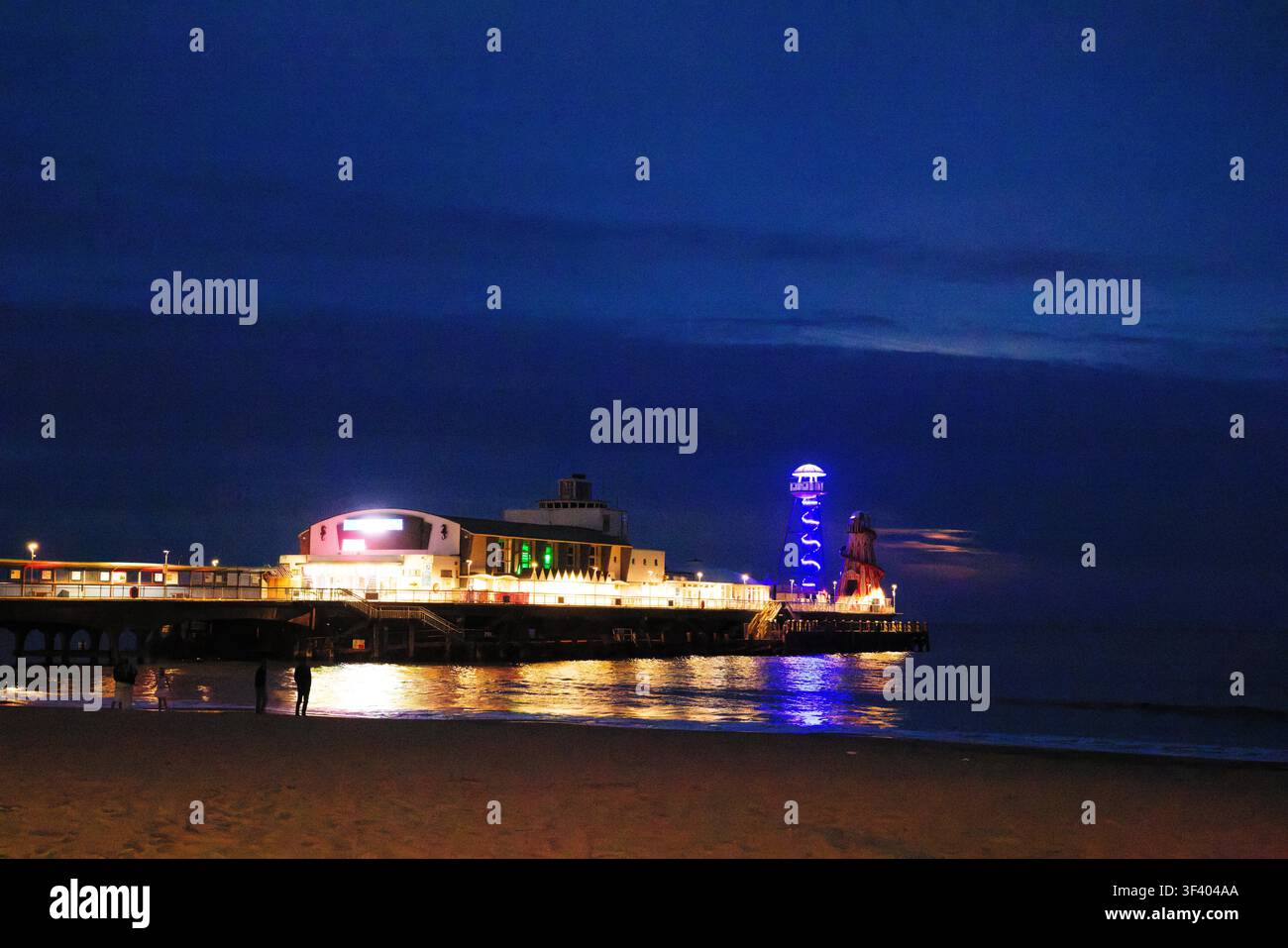 La lumière de la lune couleur cuivre se noie dans d'épais nuages bleus au crépuscule avec une jetée de plaisir brillamment éclairée sur une mer calme bleu foncé, Bournemouth, Dorset. Banque D'Images