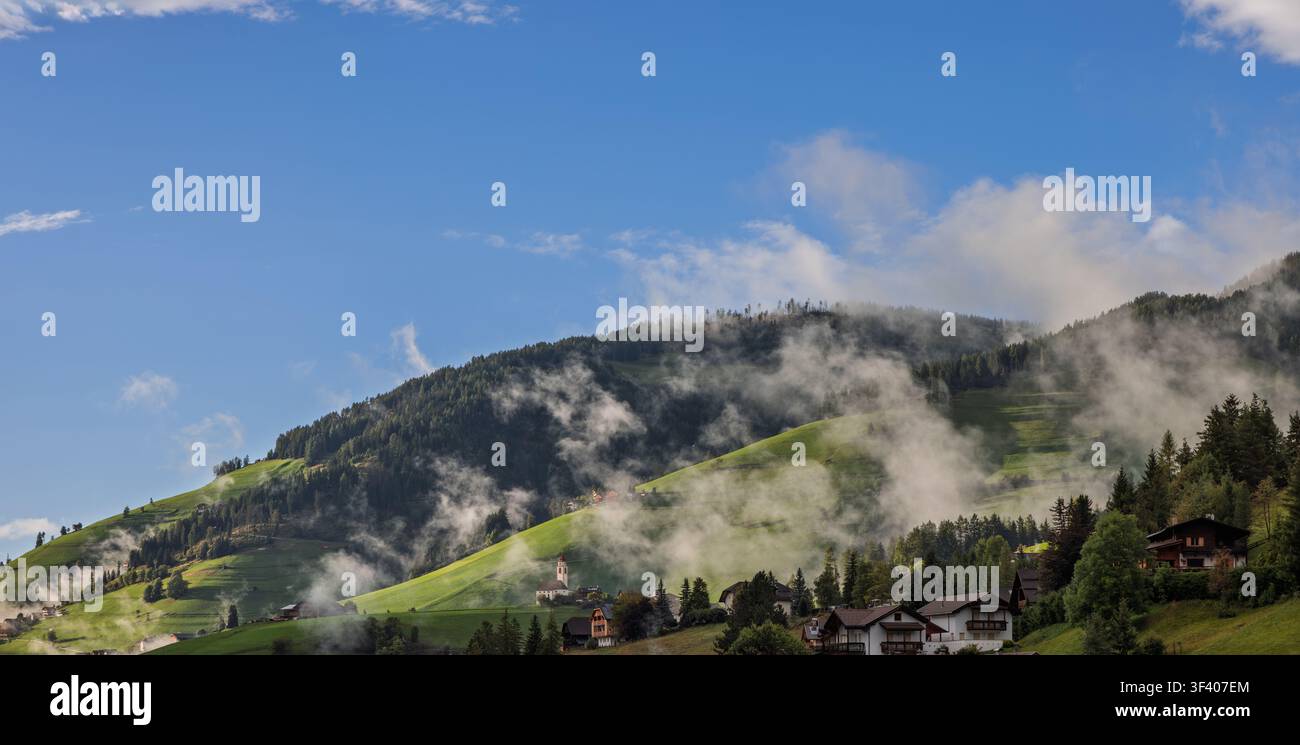 Un large panorama s'étend sur des collines vertes brumeuses, des chalets dispersés et l'église au-dessus de San Vigilio di Marebbe alors que le brouillard bas se dissipe sous le soleil. Le fo recousu Banque D'Images
