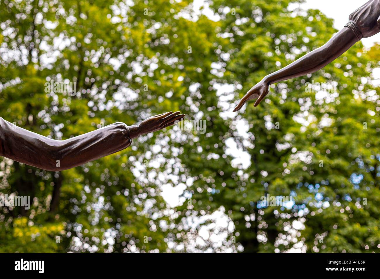 Mains sculptées en bronze se tendant l'une vers l'autre. Deux bras sculptés en bronze se tendent l'un vers l'autre devant un baldaquin légèrement flou de vert illuminé par le soleil Banque D'Images