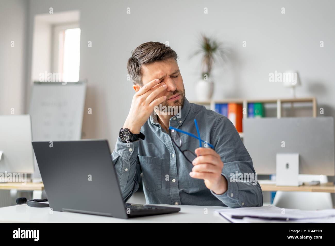 Jeune homme stressé tenant des lunettes et se frottant le front pendant qu'il travaille au bureau, concept d'affaires Banque D'Images